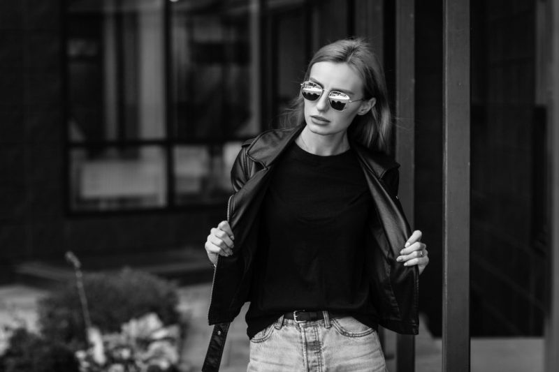 Girl wearing t-shirt, glasses and leather jacket posing against street , urban clothing style. Street photography